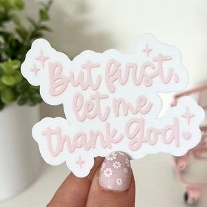 May include: A white sticker with the text "But first, let me thank God" in a soft pink color. The sticker has a white border and decorative star and heart accents. The sticker is held by a person.