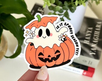Könnte beinhalten: Ein weißer Geist mit einem fröhlichen Gesicht schaut aus einem lächelnden orangefarbenen Kürbis heraus. Der Text "Is it spooky season yet?" ist in einem Kreis um den Geist und den Kürbis geschrieben.