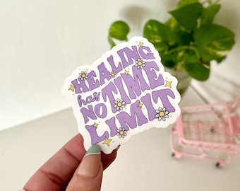 May include: Purple sticker with the text "Healing has no time limit" in a retro font. The sticker is decorated with small yellow stars and white daisies.