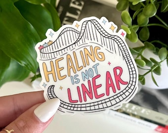 May include: A white sticker with a black and white roller coaster design. The text "Healing is not linear" is written in colorful letters on the sticker.
