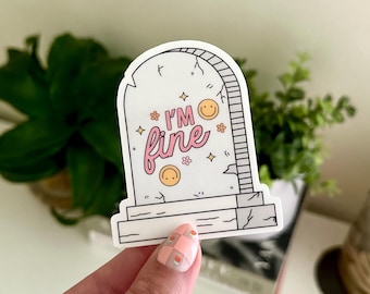 May include: White sticker with a tombstone design. The tombstone has a cracked surface and says "I'm fine" in pink lettering with a smiling face. There are small flowers and stars around the text.