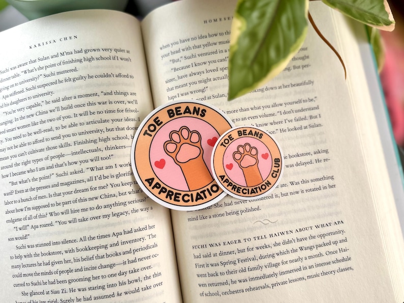 Pu&ograve; includere: Due adesivi rotondi con un'impronta di zampa e il testo "Toe Beans Appreciation Club". L'adesivo pi&ugrave; grande &egrave; arancione e rosa con un'impronta di zampa bianca. L'adesivo pi&ugrave; piccolo &egrave; bianco con un'impronta di zampa rosa.