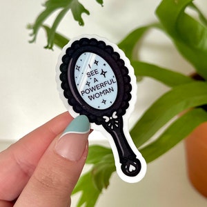 Puede incluir: Una pegatina en blanco y negro de un espejo de mano con un mango negro y un fondo blanco. El espejo refleja el texto "Veo a una mujer poderosa".