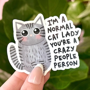 May include: A sticker featuring a gray cartoon cat with black stripes and pink cheeks. The sticker has the text "I'M A NORMAL CAT LADY YOU'RE A CRAZY PEOPLE PERSON" in black font.