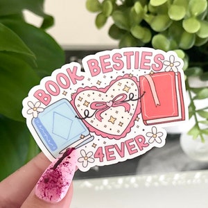 May include: A pink and white sticker with the text "Book Besties 4ever". The sticker features two books, a heart with a bow, and small white flowers.