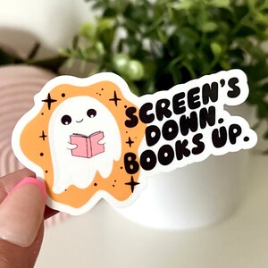 May include: A white ghost holding a pink book with the text "SCREEN'S DOWN. BOOKS UP." on a yellow background with black stars.