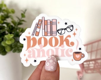 Peut inclure: Un autocollant blanc avec un design rose et marron qui dit "bookaholic". L'autocollant présente une pile de livres, une paire de lunettes, une tasse de café et des étoiles.