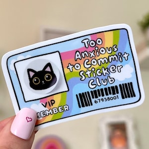 Puede incluir: Una pegatina con la cara de un gato de dibujos animados y el texto "Too Anxious to Commit Sticker Club". La pegatina tiene un fondo de arcoíris y un código de barras. El gato tiene una cara negra y ojos amarillos grandes.