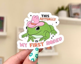 Puede incluir: Una rana verde con un sombrero de vaquero rosa con una estrella blanca. La rana está sentada sobre un fondo blanco con el texto "This is actually my first rodeo" en rosa, naranja y blanco.