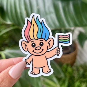 May include: A sticker featuring a cartoon troll with rainbow hair and a rainbow flag. The troll has a smiling face and is holding the flag in its right hand. The sticker has a white border.