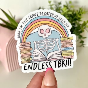Könnte beinhalten: Ein Aufkleber mit einem Skelett, das unter einem Regenbogen ein Buch liest. Das Buch ist mit "Endless TBR!!" beschriftet und der Buchrücken ist mit verschiedenen Romantik-Tropen beschriftet, darunter "Slow Burn", "Dark Romance", "Enemies to Lovers", "Grumpy Sunshine", "Spicy Fairytale", "One Bed", "Age Gap", "Shut With Plat", "Fated Mates", "Friends to Lovers", "Opposites Attract", "Midas Romance" und "Forbidden Love".