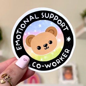 May include: A round sticker with a black background and white text that reads "Emotional Support Co-Worker". The sticker features a cartoon brown bear with a pink nose and black eyes on a rainbow background with white stars.