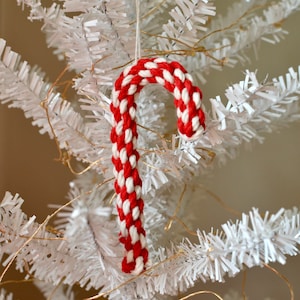 Könnte beinhalten: Ein rot-weiß geflochtener Zuckerstangen-Weihnachtsschmuck, der an einem weißen Weihnachtsbaum hängt. Die Zuckerstange besteht aus dickem, gewebtem Seil und hat ein klassisches Streifenmuster. Der Hintergrund ist verschwommen weiß.