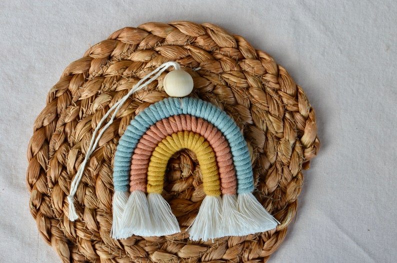 May include: A colourful rainbow wall hanging made of yarn with a wooden bead at the top. The rainbow has three colours: blue, peach, and yellow. The rainbow has white tassels at the bottom.