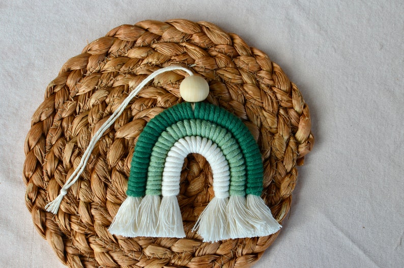 May include: A green and white ombre rainbow macrame wall hanging with white tassels. The rainbow is attached to a wooden bead.