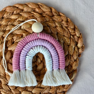 May include: A pink and white rainbow macrame wall hanging with a wooden bead at the top. The rainbow is made of cotton cord and has a fringe at the bottom.