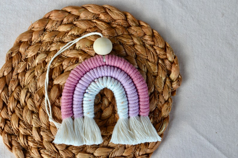 May include: A pink and white rainbow macrame wall hanging with a wooden bead at the top. The rainbow is made of cotton cord and has a fringe at the bottom.
