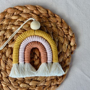 May include: A rainbow-coloured macrame wall hanging with a wooden bead at the top. The rainbow is made of yellow, pink, and orange yarn and has white fringe at the bottom.