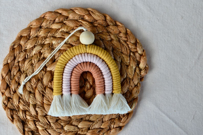May include: A rainbow-coloured macrame wall hanging with a wooden bead at the top. The rainbow is made of yellow, pink, and orange yarn and has white fringe at the bottom.