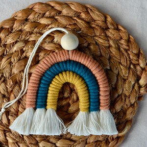 May include: A colourful rainbow wall hanging made of yarn with a wooden bead at the top. The rainbow has three stripes: peach, teal, and yellow. The rainbow has white tassels at the bottom.