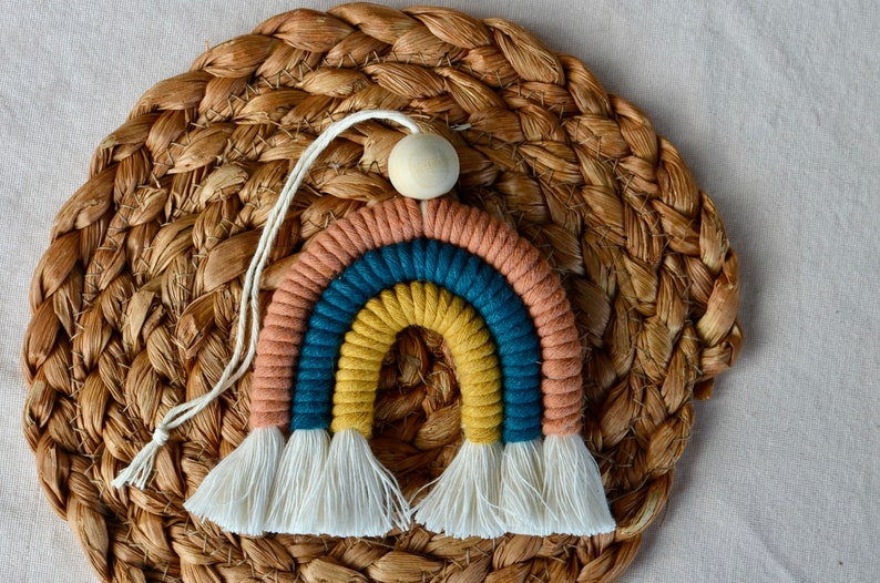 May include: A colourful rainbow wall hanging made of yarn with a wooden bead at the top. The rainbow has three stripes: peach, teal, and yellow. The rainbow has white tassels at the bottom.
