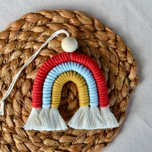 May include: A colourful rainbow macrame wall hanging with a wooden bead at the top. The rainbow is made of red, yellow, blue, and white yarn.