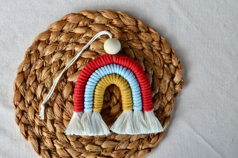 May include: A colourful rainbow macrame wall hanging with a wooden bead at the top. The rainbow is made of red, yellow, blue, and white yarn.