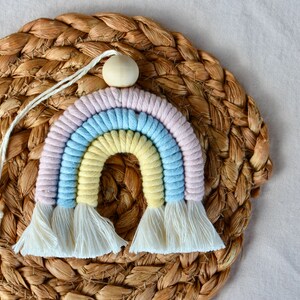 May include: A pastel rainbow wall hanging made of yarn with a wooden bead at the top. The rainbow is on a woven straw background.