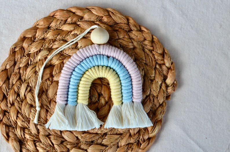 May include: A pastel rainbow wall hanging made of yarn with a wooden bead at the top. The rainbow is on a woven straw background.