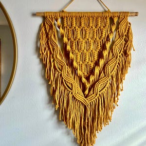 May include: Mustard yellow macrame wall hanging. The handmade textile art piece features intricate knotting patterns and a fringed bottom. It is suspended from a wooden dowel and a hanging cord, adding a bohemian touch to home decor.