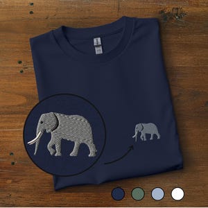 May include: A navy blue t-shirt featuring a small embroidered grey elephant on the chest. A circular inset shows a magnified view of the elephant embroidery, which depicts a walking elephant with white tusks. Four small coloured circles appear below.