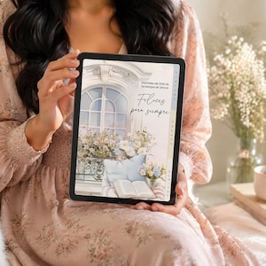 Pode incluir: Um tablet digital exibindo uma ilustração em aquarela de uma varanda com flores, uma almofada e um livro aberto. O tablet tem o texto "Felices para siempre". A pessoa segurando o tablet está usando um vestido floral.
