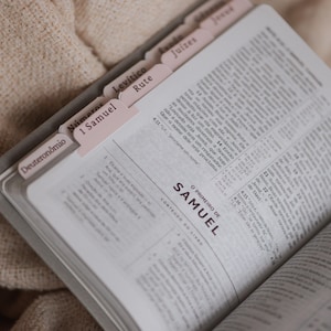 May include: An open Bible with pink tab dividers. The text is in Portuguese, with the title "1 Samuel" visible. The book is resting on a textured, beige surface.