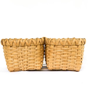 May include: Two small, square woven baskets made of light brown natural fibers. The baskets have a tightly woven pattern and a decorative border around the top. They are side-by-side on a white background.