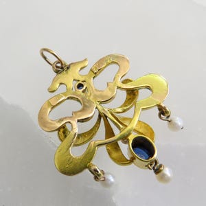 May include: A gold pendant with an ornate, swirling design. It features two oval, deep blue gemstones and three small, dangling pearls. The pendant has a loop at the top for a chain.