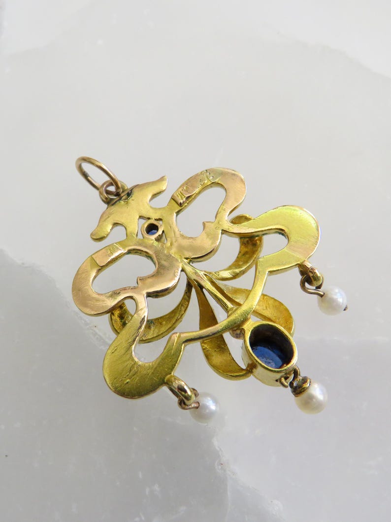 May include: A gold pendant with an ornate, swirling design. It features two oval, deep blue gemstones and three small, dangling pearls. The pendant has a loop at the top for a chain.
