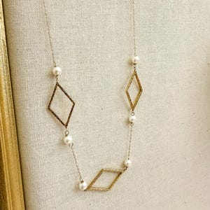May include: A delicate gold-toned necklace featuring three diamond-shaped pendants and several small, round pearls. The necklace hangs on a fine chain, creating an elegant and minimalist design. The background is a neutral, textured fabric.
