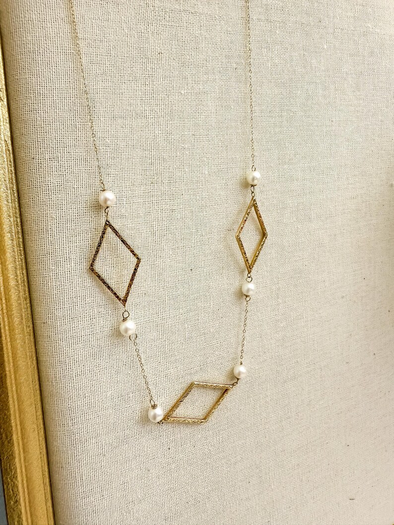 May include: A delicate gold-toned necklace featuring three diamond-shaped pendants and several small, round pearls. The necklace hangs on a fine chain, creating an elegant and minimalist design. The background is a neutral, textured fabric.