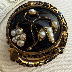 May include: A vintage round brooch with a black enamel background and gold filigree border. The brooch features a floral design with faux pearls and a small diamond accent. The outer edge has a decorative gold and black pattern.