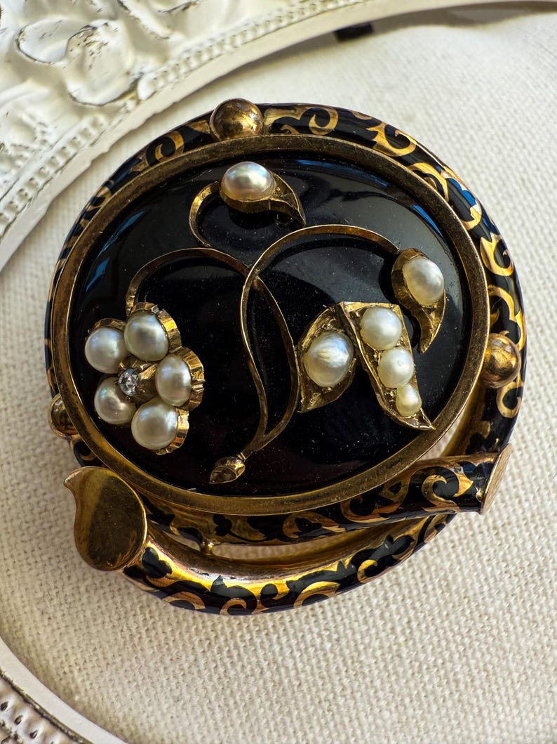 May include: A vintage round brooch with a black enamel background and gold filigree border. The brooch features a floral design with faux pearls and a small diamond accent. The outer edge has a decorative gold and black pattern.