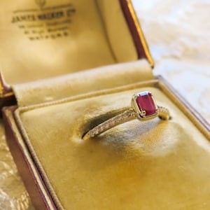 May include: A gold ring featuring a rectangular red gemstone set in the center, flanked by small clear stones embedded along the band. The ring is presented inside a vintage-style jewelry box with a cream-colored velvet interior lining.