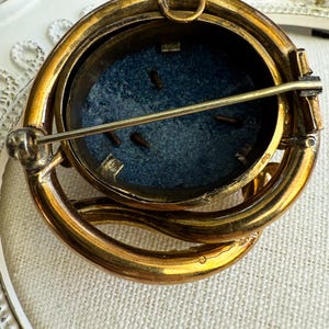 May include: Vintage gold-toned brooch with an oval, dark blue center. The brooch has a detailed, layered design with a pin closure on the back. The brooch's design is intricate, with a loop at the top for potential use as a pendant.
