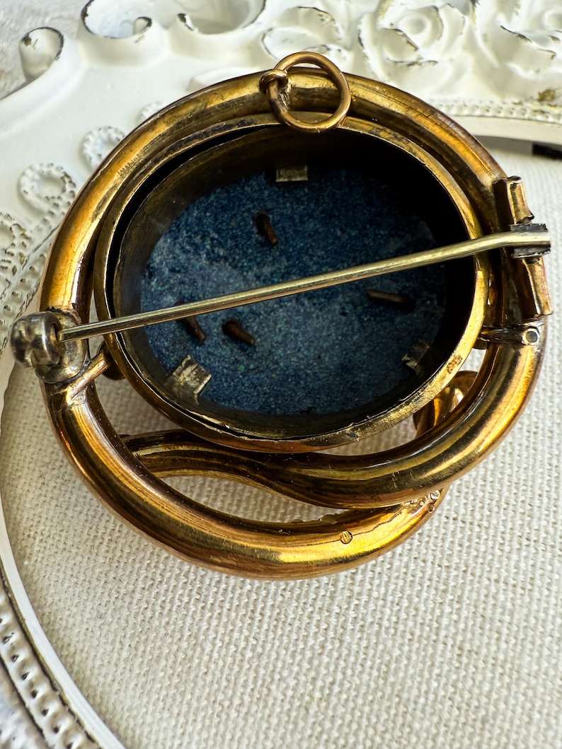 May include: Vintage gold-toned brooch with an oval, dark blue center. The brooch has a detailed, layered design with a pin closure on the back. The brooch's design is intricate, with a loop at the top for potential use as a pendant.