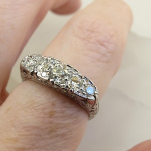 Op de afbeelding: Een zilveren ring met meerdere kleine, ronde diamanten in een rij. De ring zit om een vinger en de band heeft een sierlijk, gegraveerd ontwerp. De diamanten schitteren en de ring lijkt vintage of antiek.