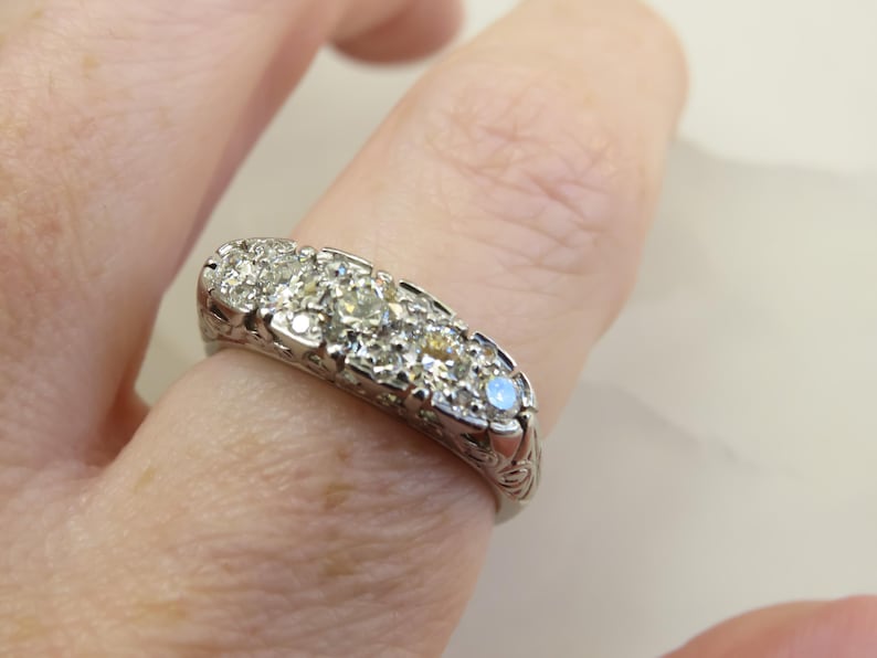 Op de afbeelding: Een zilveren ring met meerdere kleine, ronde diamanten in een rij. De ring zit om een vinger en de band heeft een sierlijk, gegraveerd ontwerp. De diamanten schitteren en de ring lijkt vintage of antiek.