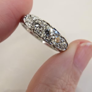 Op de afbeelding: Een zilveren ring met een rij sprankelende, heldere edelstenen. De ring wordt tussen twee vingers gehouden, wat het ingewikkelde ontwerp en de schittering van de stenen benadrukt. De zetting van de ring is gedetailleerd.