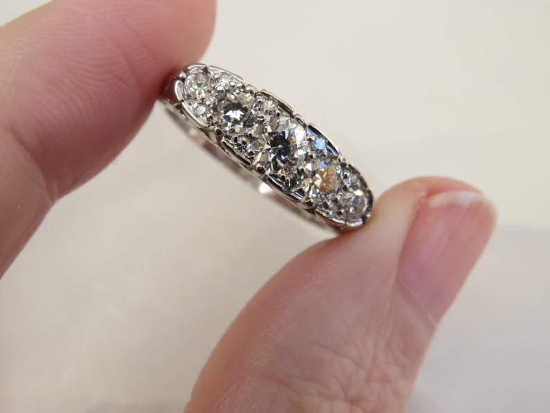 Op de afbeelding: Een zilveren ring met een rij sprankelende, heldere edelstenen. De ring wordt tussen twee vingers gehouden, wat het ingewikkelde ontwerp en de schittering van de stenen benadrukt. De zetting van de ring is gedetailleerd.