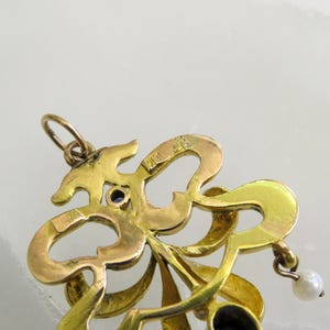 May include: A gold pendant with an elaborate, swirling design. It has a small blue gemstone and a dangling pearl. The intricate details and vintage style suggest a piece of fine jewellery, possibly antique.