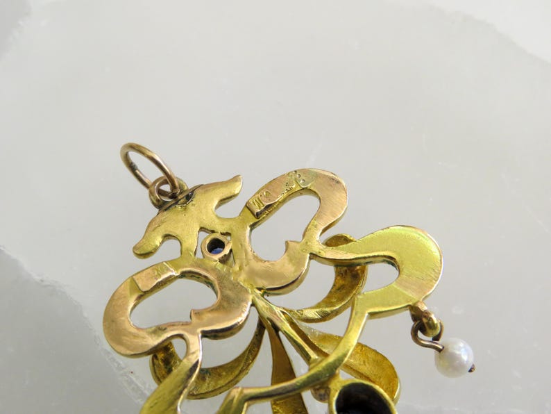 May include: A gold pendant with an elaborate, swirling design. It has a small blue gemstone and a dangling pearl. The intricate details and vintage style suggest a piece of fine jewellery, possibly antique.