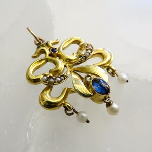 May include: A gold pendant with a floral design. It features an oval blue gemstone, small pearls, and a small dark blue gemstone. Three small pearls dangle from the bottom. The pendant is an example of fine jewellery.